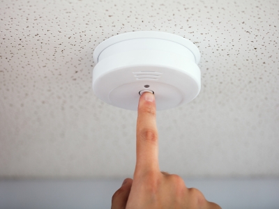 Person testing a smoke detector on the ceiling, emphasizing the importance of regular maintenance for home safety and functionality.