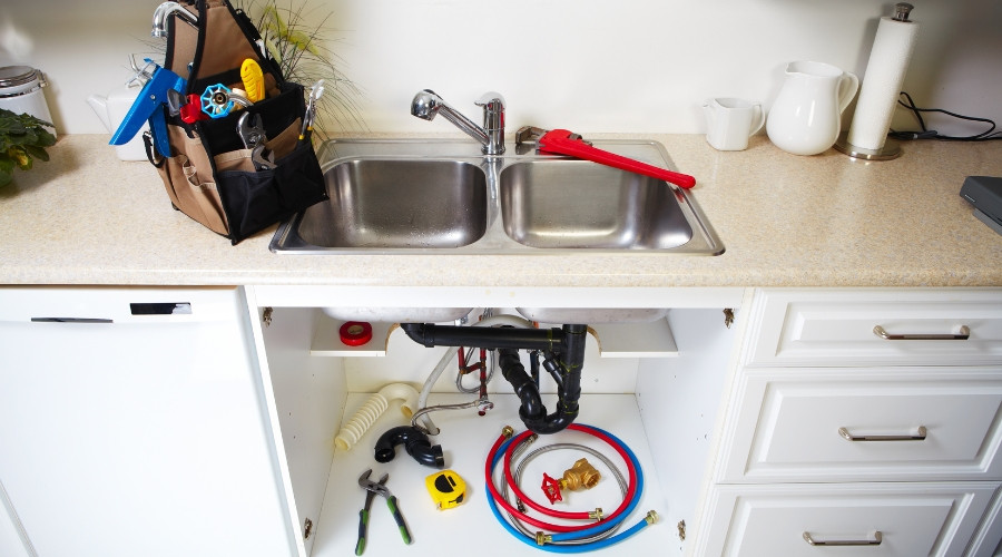 Kitchen sink with plumbing tools, including a wrench and measuring tape, under the counter, emphasizing plumbing maintenance importance.